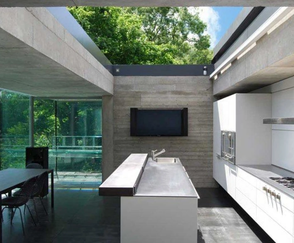 A modern kitchen features a minimalist design with concrete and glass elements. It includes sleek white cabinets, a stainless steel countertop, and a built-in oven. Large windows allow ample natural light and provide a view of greenery outside. A dining area with black chairs is visible in the background.