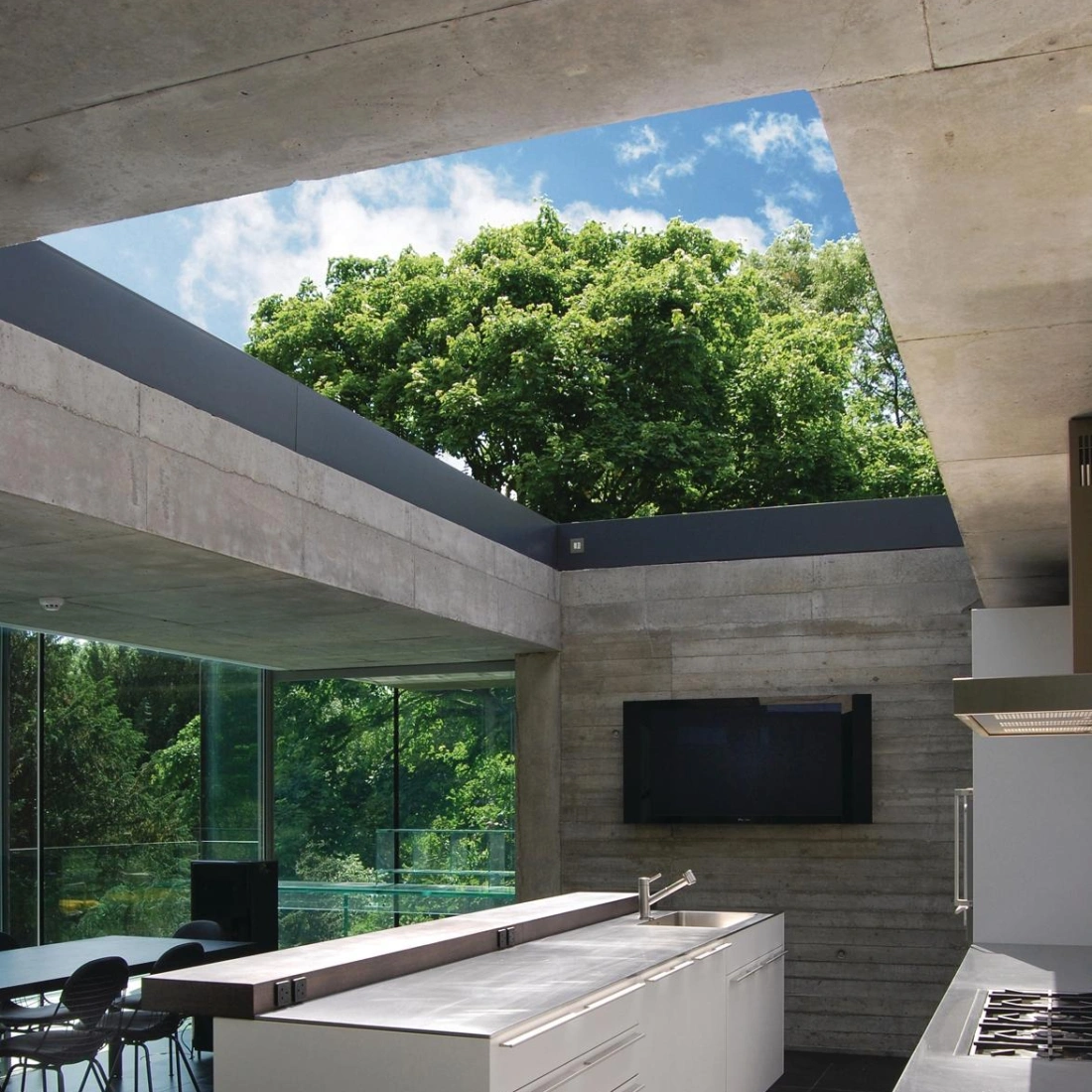Modern kitchen with a sleek white island and built-in appliances. Large windows provide a view of lush greenery outside. Minimalist design features concrete walls and dark flooring. Natural light floods the space, creating an airy atmosphere.
