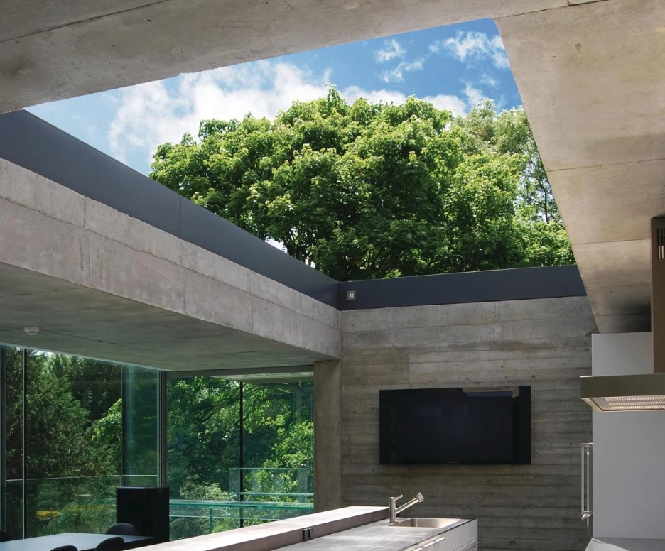 A modern kitchen features a sleek white island with drawers and a stainless steel sink, surrounded by large glass windows that overlook lush greenery. The ceiling is made of concrete, allowing natural light to flood the space. A wall-mounted television is visible in the background.