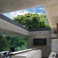Cocina moderna con diseño minimalista, muebles blancos y encimera de madera. Amplios ventanales permiten la entrada de luz natural y ofrecen vistas de un árbol exterior. Suelo de cerámica oscura.