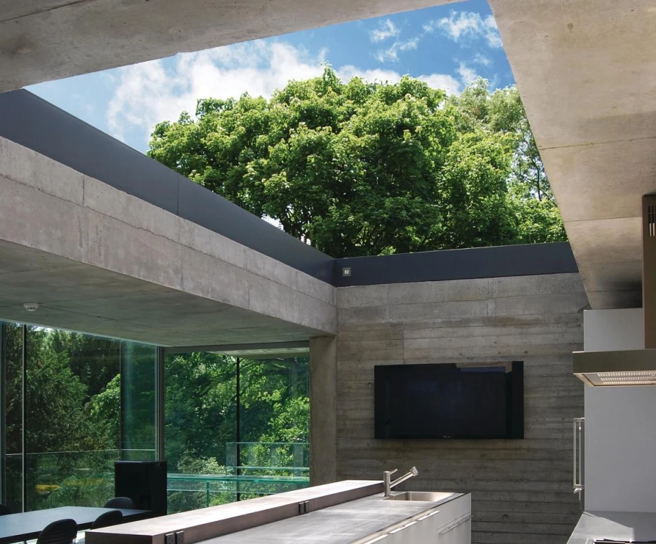Cocina moderna con una isla central blanca, paredes de concreto y grandes ventanales que ofrecen vistas a un árbol verde. El espacio tiene un diseño minimalista y contemporáneo.