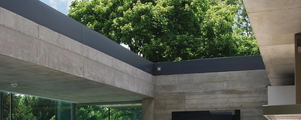 A modern kitchen features a sleek white island with drawers and a stainless steel sink, surrounded by large glass windows that overlook lush greenery. The ceiling is made of concrete, allowing natural light to flood the space. A wall-mounted television is visible in the background.