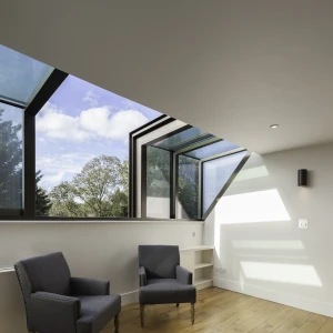 Two gray upholstered chairs are positioned in a well-lit room with large modern windows showcasing a blue sky and trees outside. Light wooden flooring complements the space, which has minimalist decor and built-in shelving.