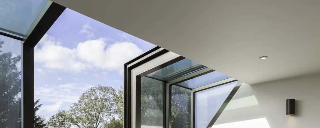 Espacio interior iluminado con dos sillones grises frente a una ventana grande con vidrio azul. El suelo es de madera clara y hay estanterías en la pared. Se pueden ver árboles y un cielo parcialmente nublado a través de la ventana.