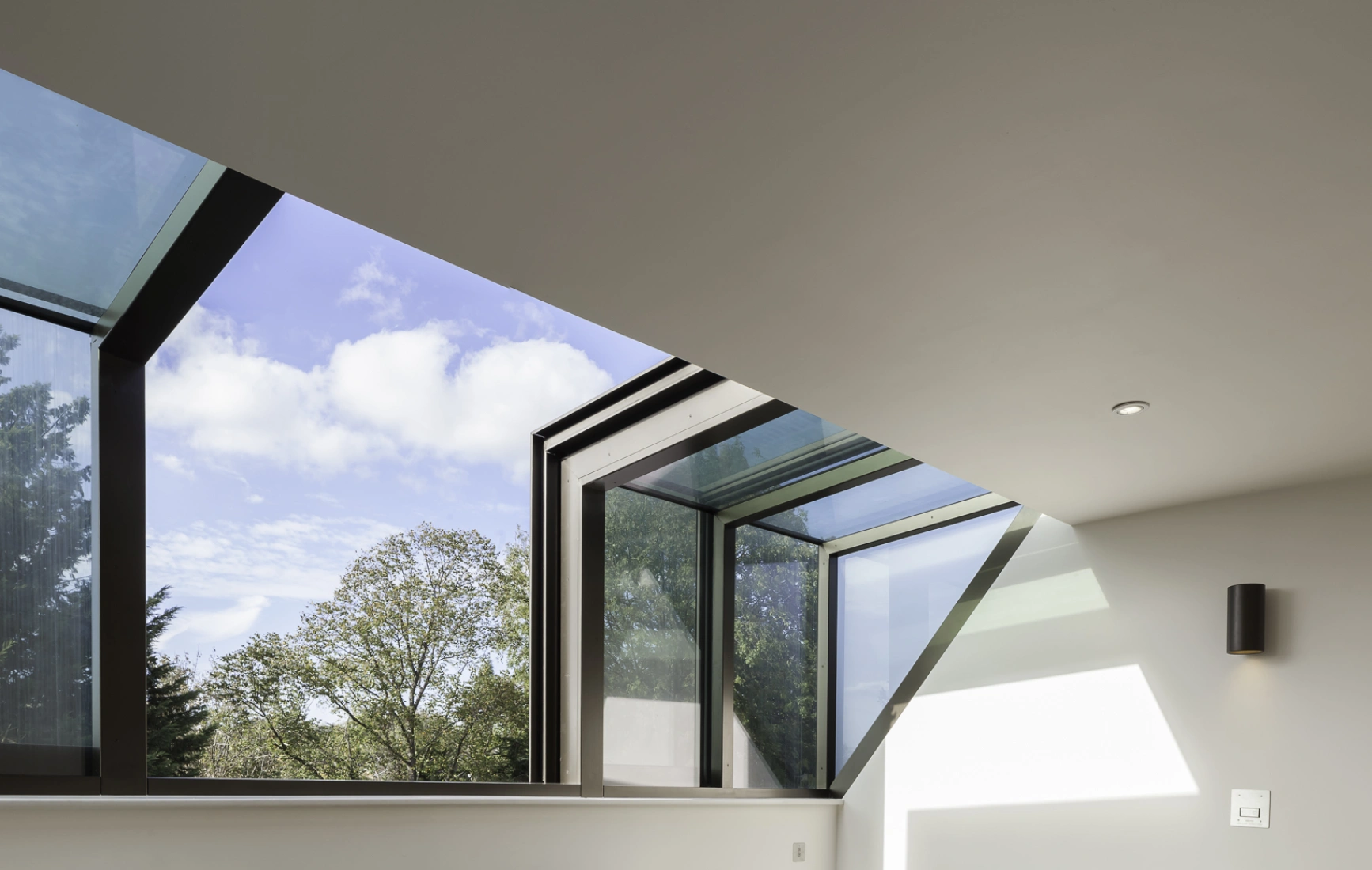 Two gray upholstered chairs are positioned in a well-lit room with large modern windows showcasing a blue sky and trees outside. Light wooden flooring complements the space, which has minimalist decor and built-in shelving.