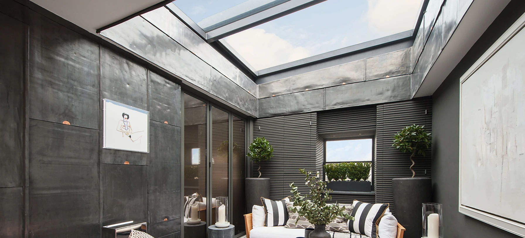 A modern sunroom featuring a sleek design with large skylights. Two sofas with white cushions and black decorative pillows are arranged around a round coffee table. The walls are a blend of dark materials, and there are potted plants and decorative elements enhancing the space. Natural light fills the room, creating a warm ambiance.