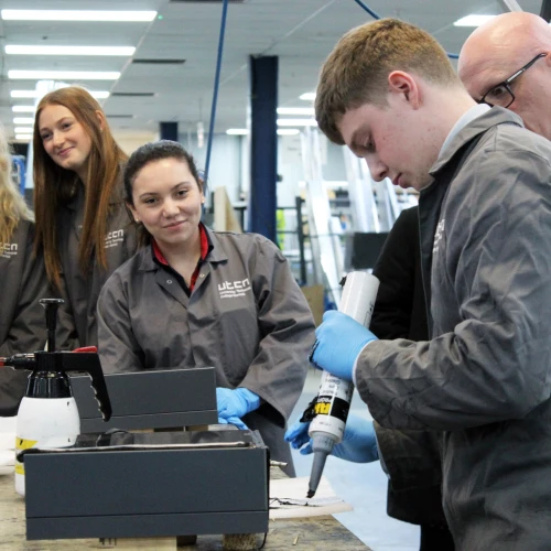 Students in lab coats gather around a workbench as one of them applies adhesive from a tube onto a surface, while others observe with interest.