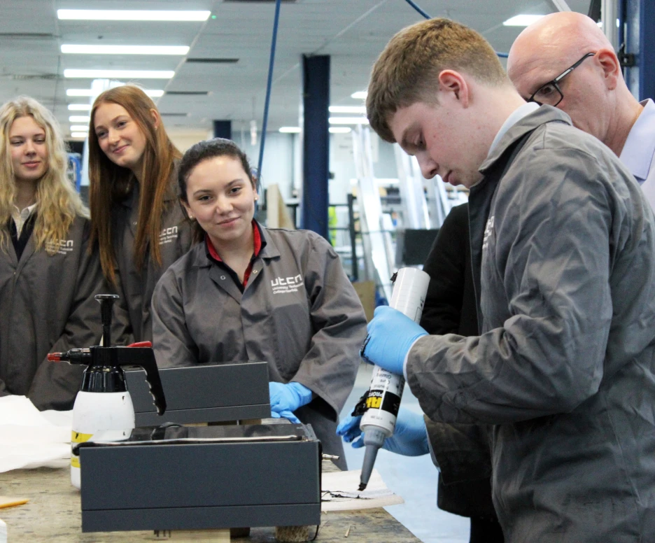 A group of students watches as a young man applies a sealant from a tube onto a project at a workbench. All participants are wearing gray work coats, and the environment appears to be a workshop or classroom setting.