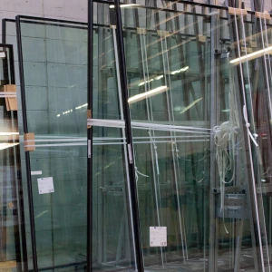 Several large glass panels are stored upright in a warehouse. The panels are partially covered with protective materials and are arranged beside wooden supports. Bright lights illuminate the area, reflecting off the glass surfaces.