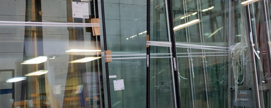 Several large glass panels are stored upright in a warehouse. The panels are partially covered with protective materials and are arranged beside wooden supports. Bright lights illuminate the area, reflecting off the glass surfaces.