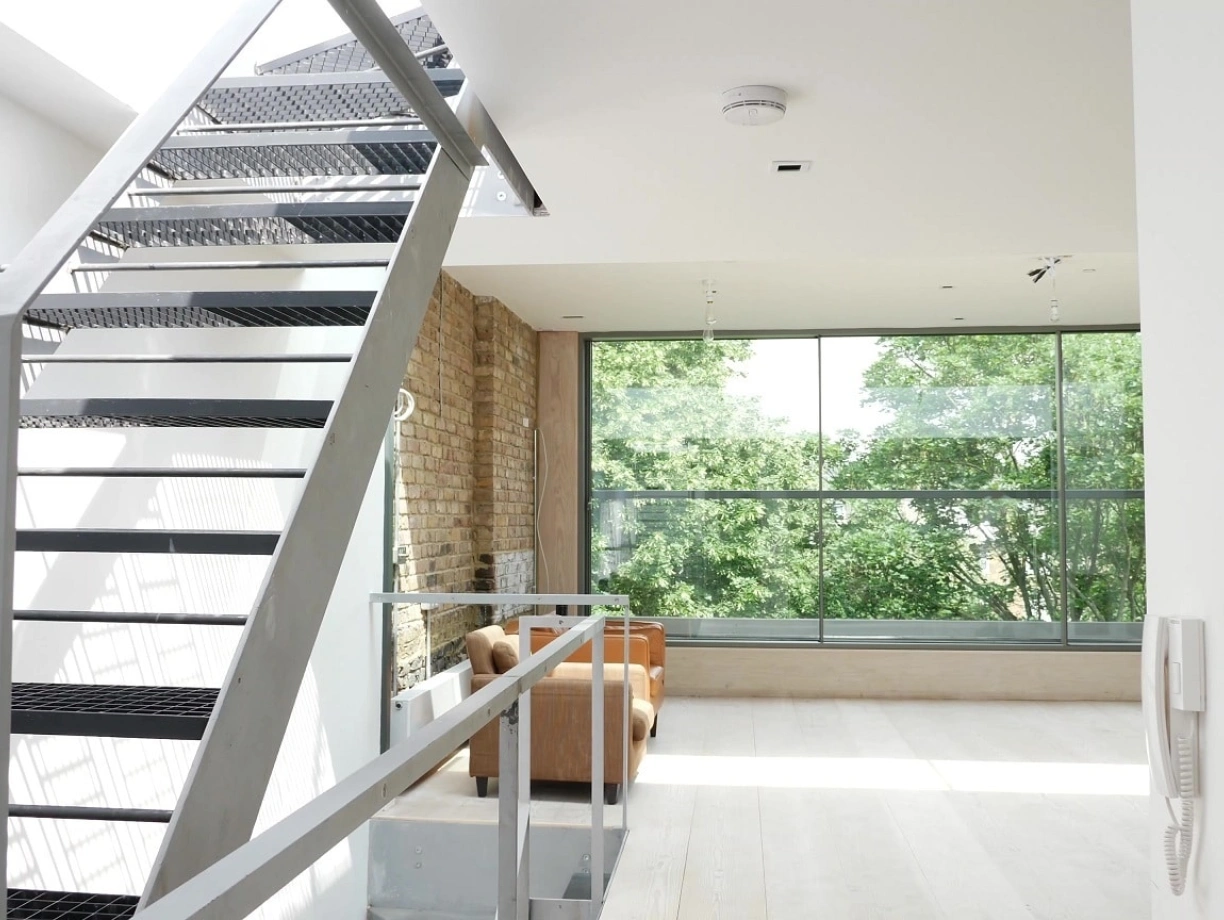A modern interior featuring a metal staircase leading to an upper level. Large windows showcase trees outside, and a cozy seating area is visible. Natural light floods the space, highlighting the minimalist design.
