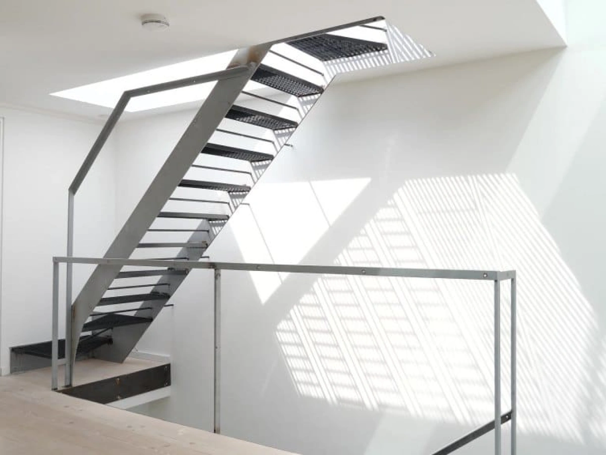 A modern, open staircase with a sleek metal structure and an upper landing. Natural light filters through a skylight, casting shadows on the white walls and wooden floor.