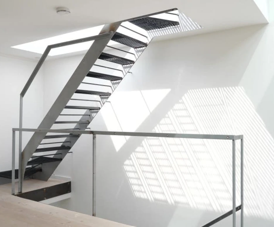 A modern, open staircase with a sleek metal structure and an upper landing. Natural light filters through a skylight, casting shadows on the white walls and wooden floor.