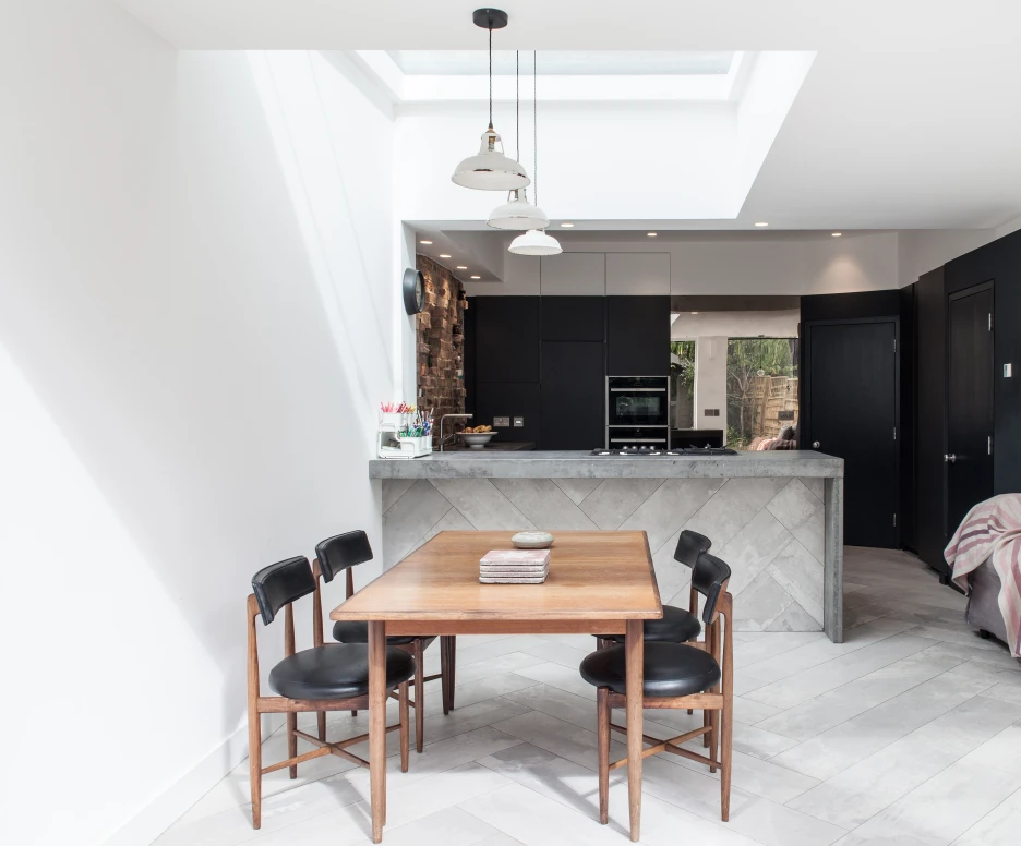 A modern kitchen and dining area with a wooden dining table surrounded by black chairs. The kitchen features dark cabinetry and a stone countertop, with natural light streaming in from a skylight above. A gray sofa with a striped throw is in the corner.