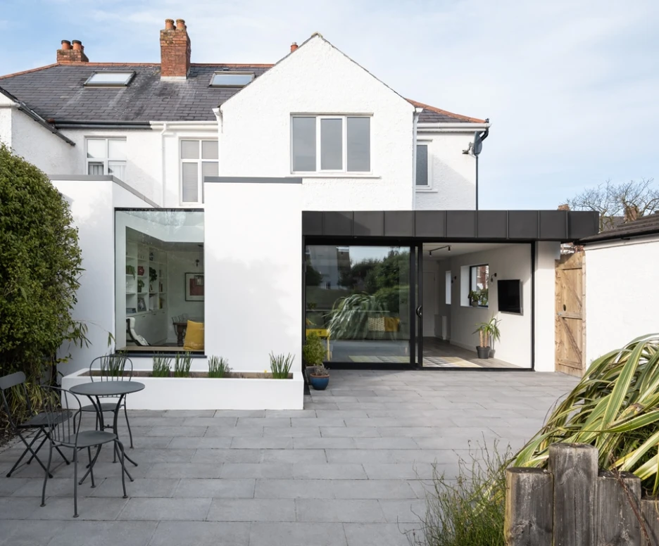 A modern home extension features large glass windows, blending sleek design with a cozy outdoor seating area. The patio is paved with gray tiles and includes a small table and chairs, surrounded by greenery.
