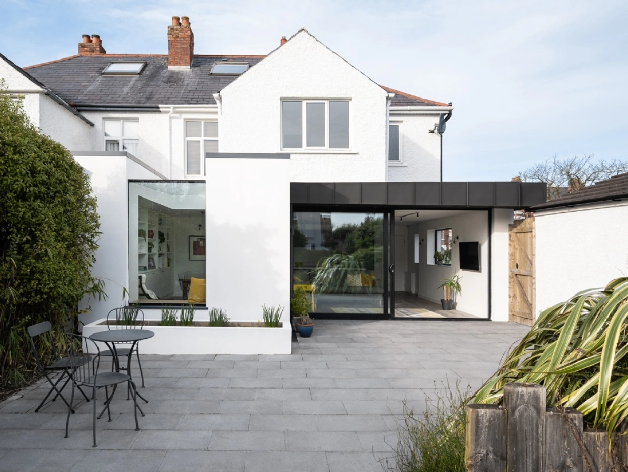 A modern home extension features large glass windows, blending sleek design with a cozy outdoor seating area. The patio is paved with gray tiles and includes a small table and chairs, surrounded by greenery.