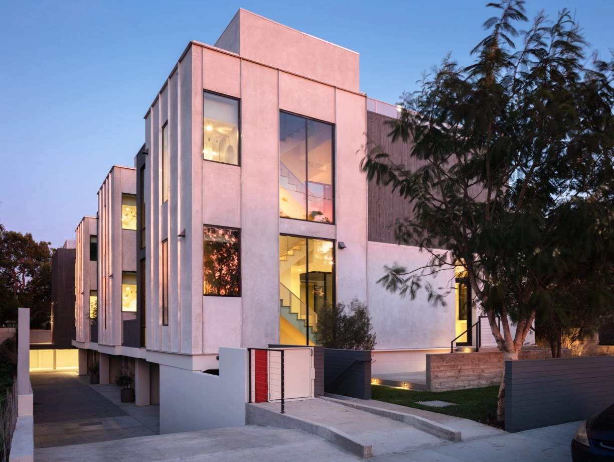 Modern multi-story building with large windows and a mix of textured concrete and smooth facades. Soft evening light highlights the structure, surrounded by landscaped greenery and a driveway.