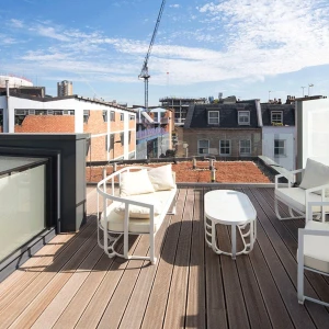 A rooftop terrace featuring white lounge chairs and a small table, with a view of adjacent buildings and cranes against a blue sky with scattered clouds.