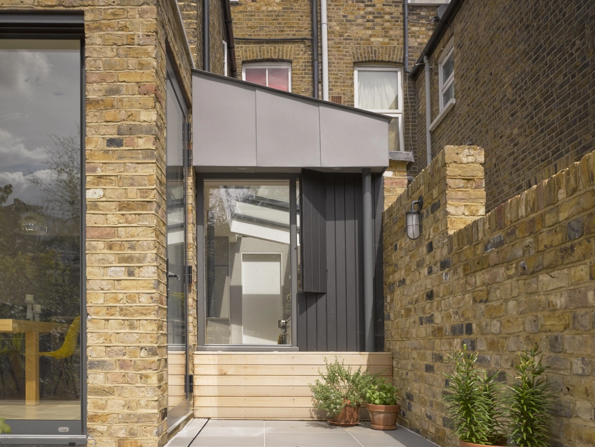 Une extension moderne de maison avec des murs en briques et une toiture inclinée, entourée de dalles en pierre. Un petit pot de plante verte est visible à côté de l’entrée. Les fenêtres sont grandes et la lumière naturelle entre abondamment.