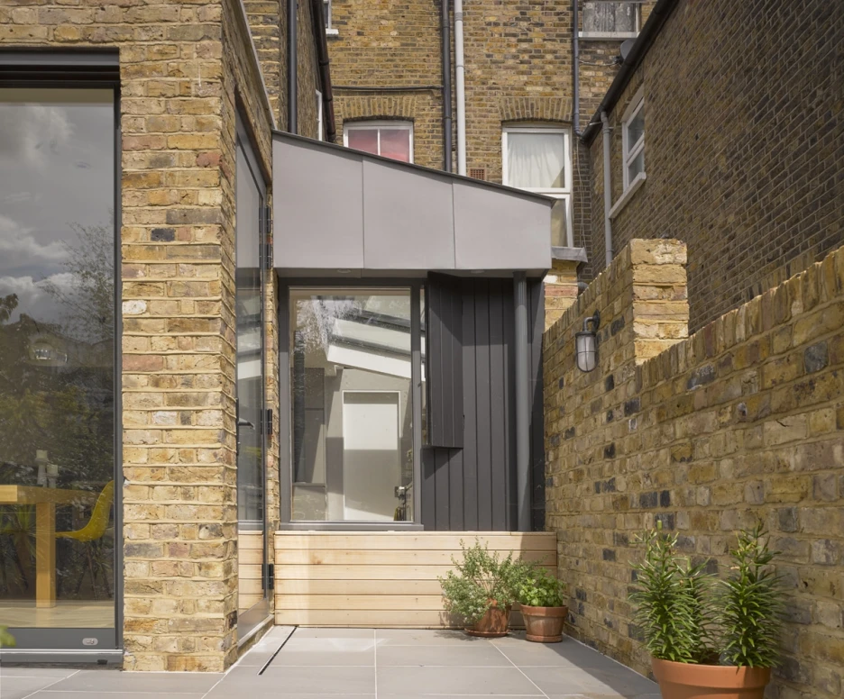 Une extension moderne de maison avec des murs en briques et une toiture inclinée, entourée de dalles en pierre. Un petit pot de plante verte est visible à côté de l’entrée. Les fenêtres sont grandes et la lumière naturelle entre abondamment.