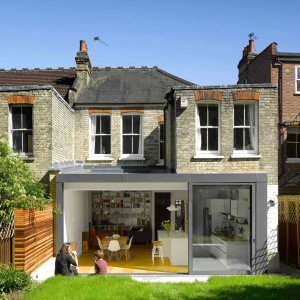 A modern glass extension leads to a cozy garden, featuring a couple sitting on the grass. The back of a traditional brick house contrasts with the sleek, contemporary design of the extension. Clear blue skies enhance the cheerful atmosphere.