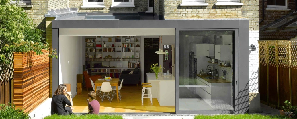 A modern glass extension leads to a cozy garden, featuring a couple sitting on the grass. The back of a traditional brick house contrasts with the sleek, contemporary design of the extension. Clear blue skies enhance the cheerful atmosphere.