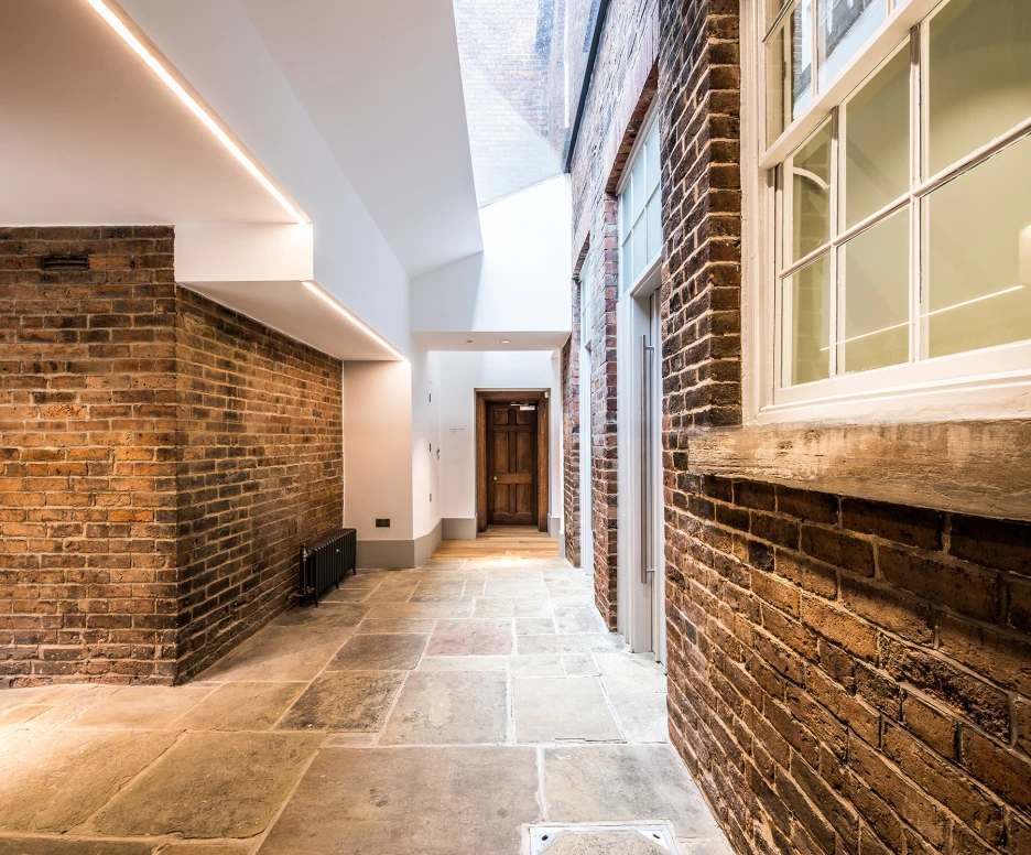 A modern hallway with a mix of brick and smooth walls, featuring a stone floor and a skylight above. A wooden door is visible at the end of the corridor, and large windows allow natural light to enter.