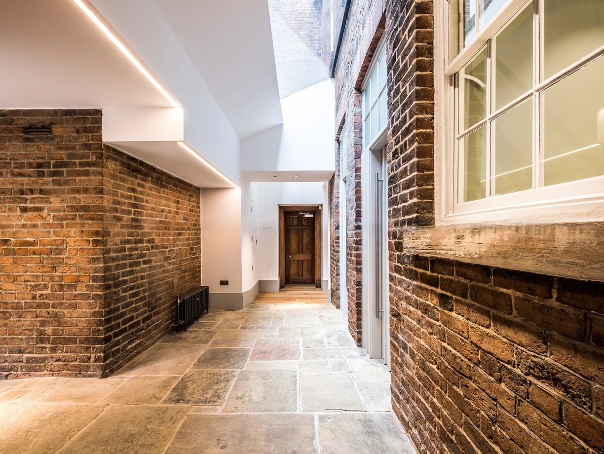 A modern hallway with a mix of brick and smooth walls, featuring a stone floor and a skylight above. A wooden door is visible at the end of the corridor, and large windows allow natural light to enter.