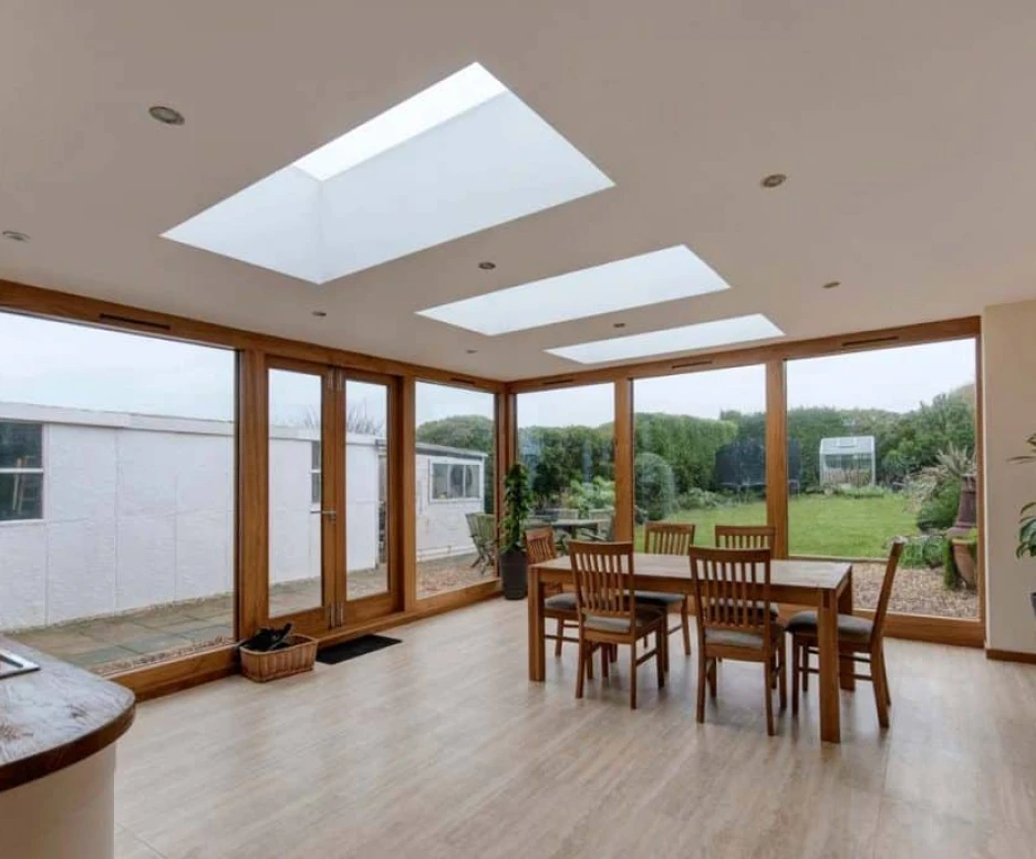 A modern dining area features a wooden table surrounded by chairs, large skylights above, and sliding glass doors leading to a green outdoor space. Light-colored walls and wooden flooring enhance the airy ambiance.