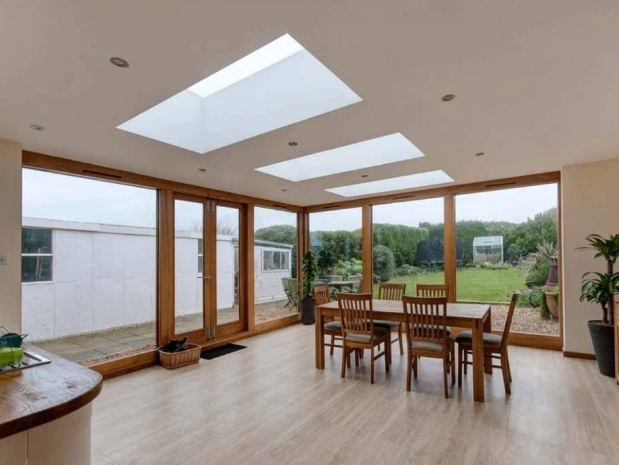 A modern dining area features a wooden table surrounded by chairs, large skylights above, and sliding glass doors leading to a green outdoor space. Light-colored walls and wooden flooring enhance the airy ambiance.