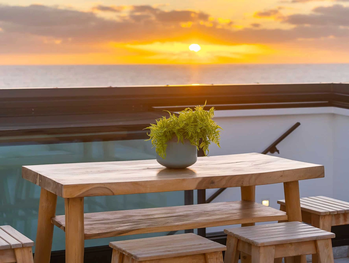 A wooden table set with stools on a balcony, featuring a potted plant in the center, against a backdrop of a vibrant sunset over the ocean.