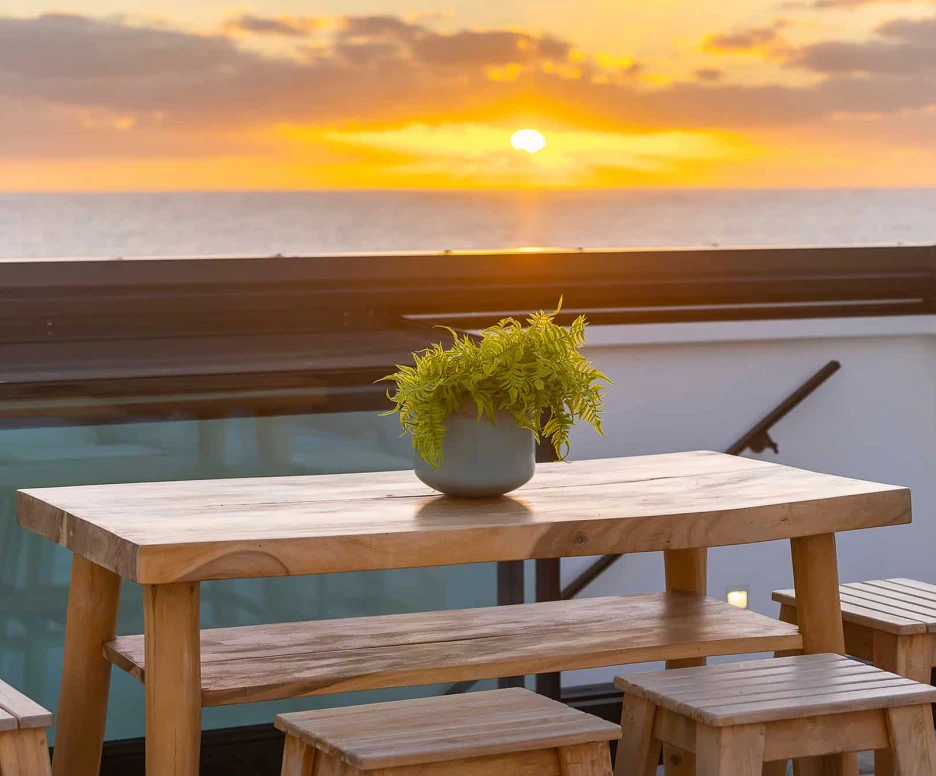 A wooden table set with stools on a balcony, featuring a potted plant in the center, against a backdrop of a vibrant sunset over the ocean.