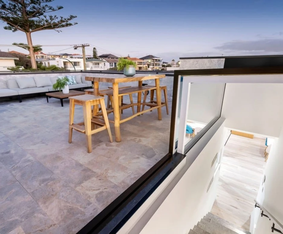 Terraza moderna con muebles de exterior, una mesa alta de madera y un sofá. Se observa un entorno residencial con casas y árboles de fondo, y escaleras que conducen a un espacio interior. La luz del atardecer crea una atmósfera acogedora.