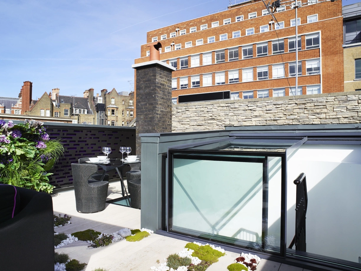 A modern rooftop terrace features a glass canopy, a table set for two with black chairs, and greenery among stone and wooden elements. In the background, a multi-story brick building is visible against a clear blue sky.