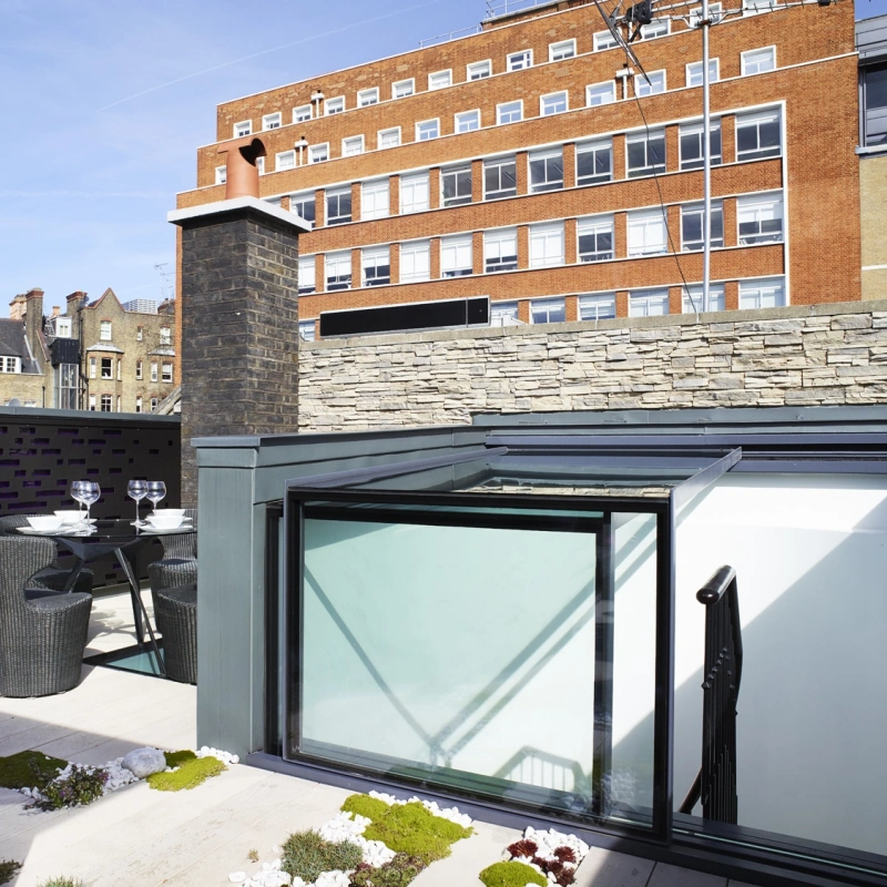 A modern rooftop terrace features a glass enclosure, a small table set for two, and decorative plants. A brick building and urban skyline are visible in the background.