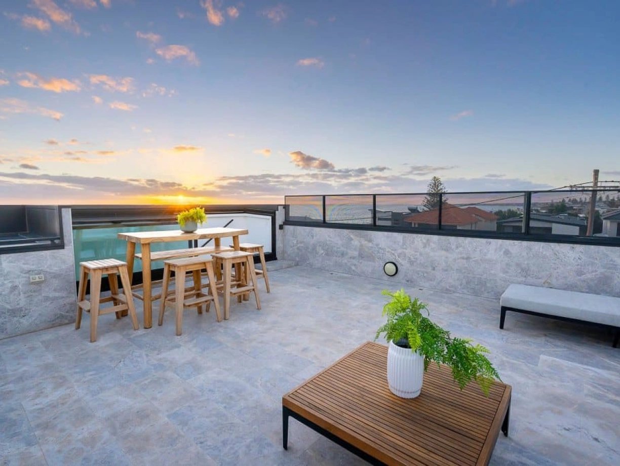 A modern rooftop terrace features a wooden dining set with stools, a potted plant, and a coffee table. The sky is filled with vibrant hues of orange and pink as the sun sets in the background, casting a warm glow over the area.