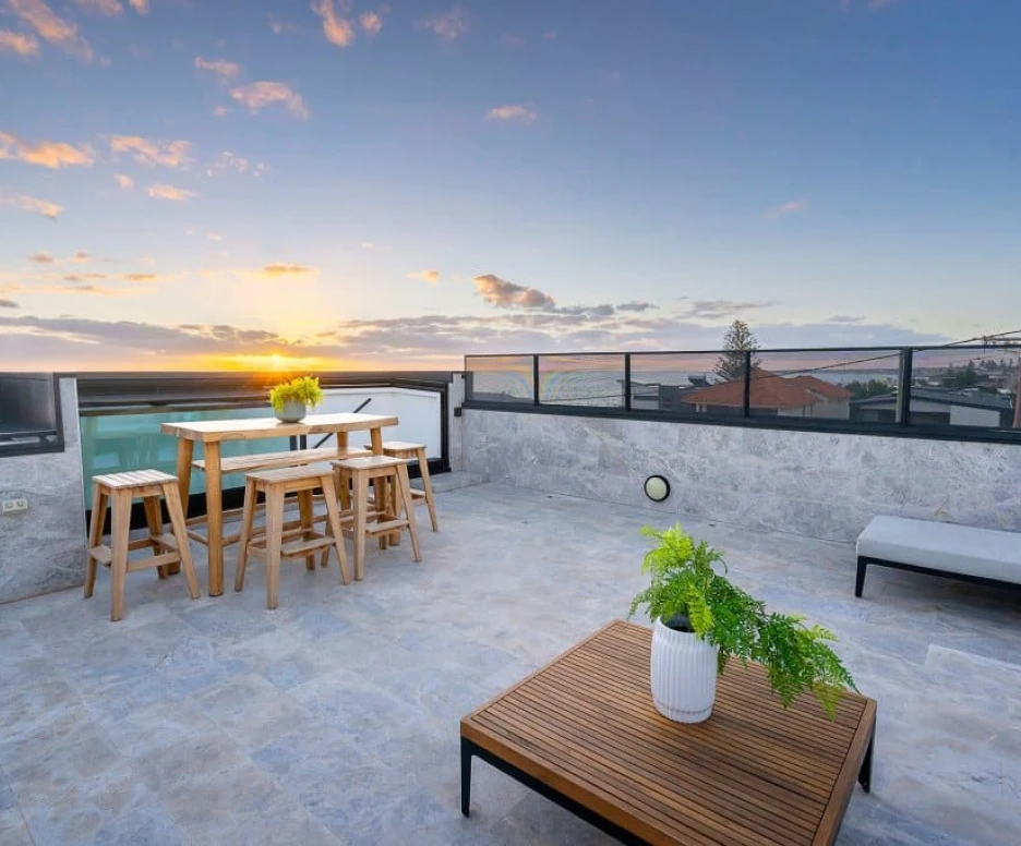 A rooftop terrace features a wooden dining table and chairs, a low wooden coffee table, and a potted plant. The scene is set against a vibrant sunset, illuminating the sky with shades of orange and pink. Surrounding buildings and a swimming pool are visible in the background.