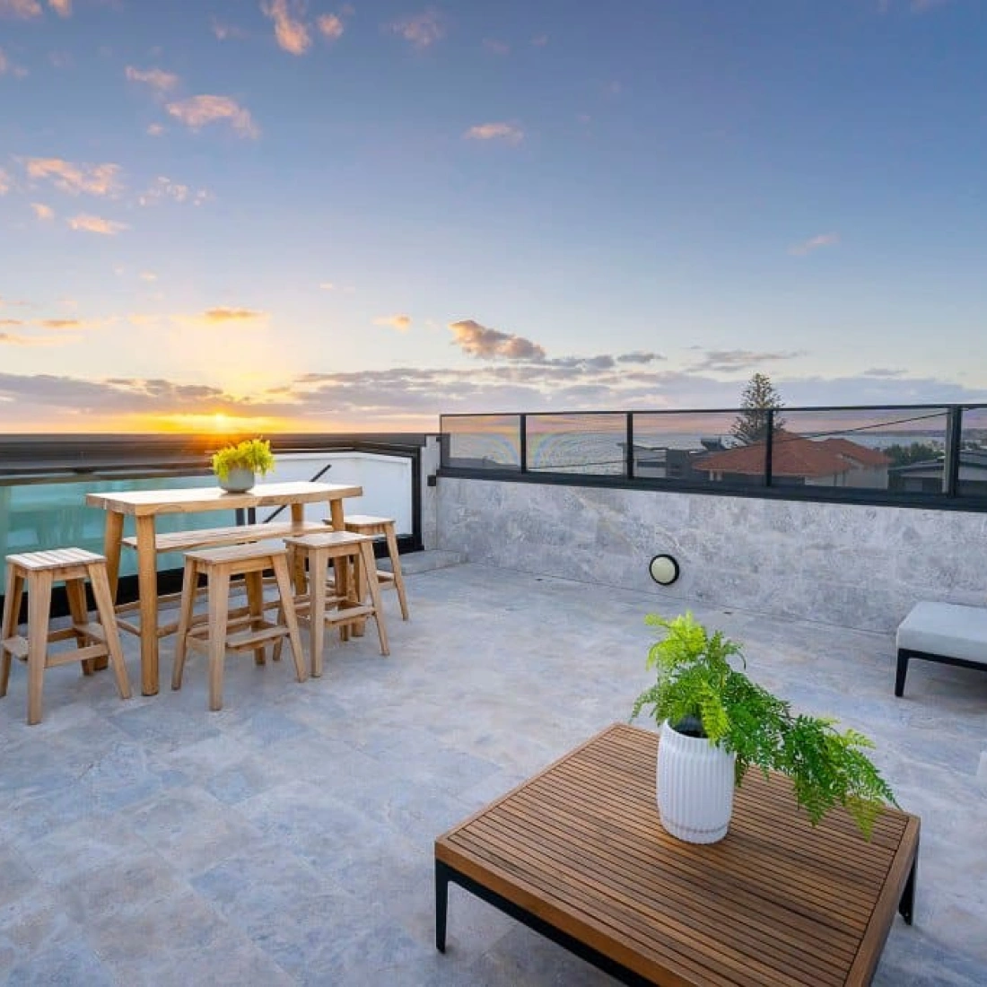 Terraza moderna con una mesa de madera y sillas, iluminada por un atardecer. Hay un sofá y una planta en una maceta blanca. El cielo muestra tonos naranjas y rosados.