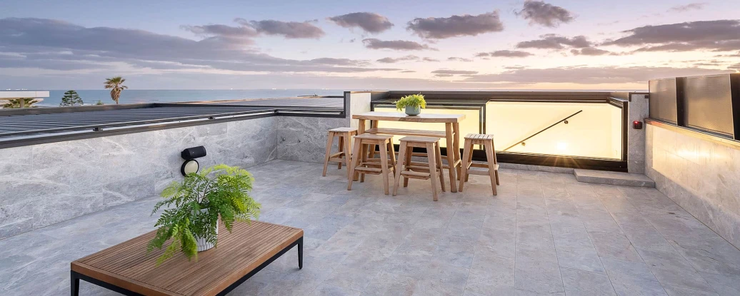 A modern rooftop terrace with a wooden table and stools, surrounded by potted plants. The area features stone flooring and has a view of a sunset sky with clouds and palm trees in the distance.