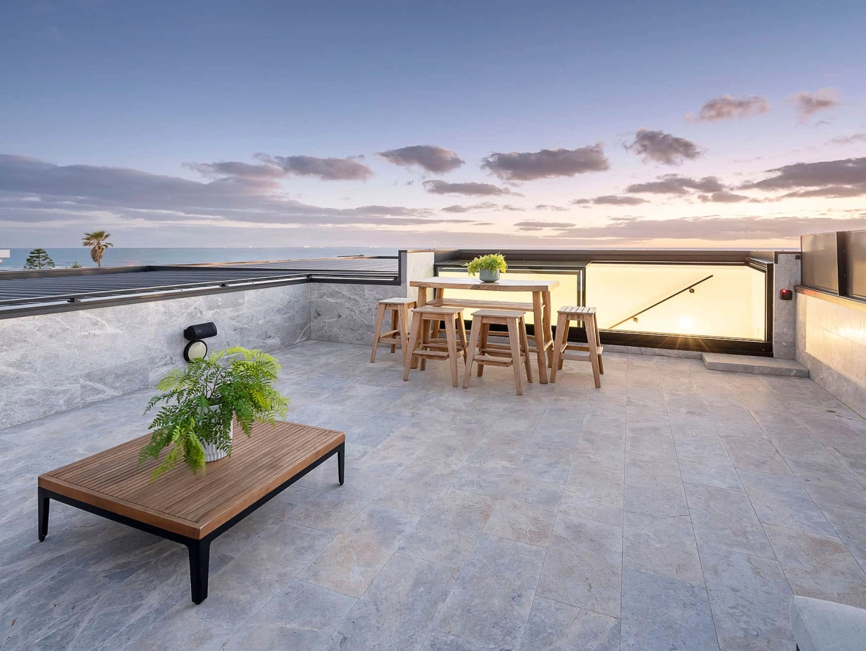 A modern rooftop terrace with a wooden table and stools, surrounded by potted plants. The area features stone flooring and has a view of a sunset sky with clouds and palm trees in the distance.