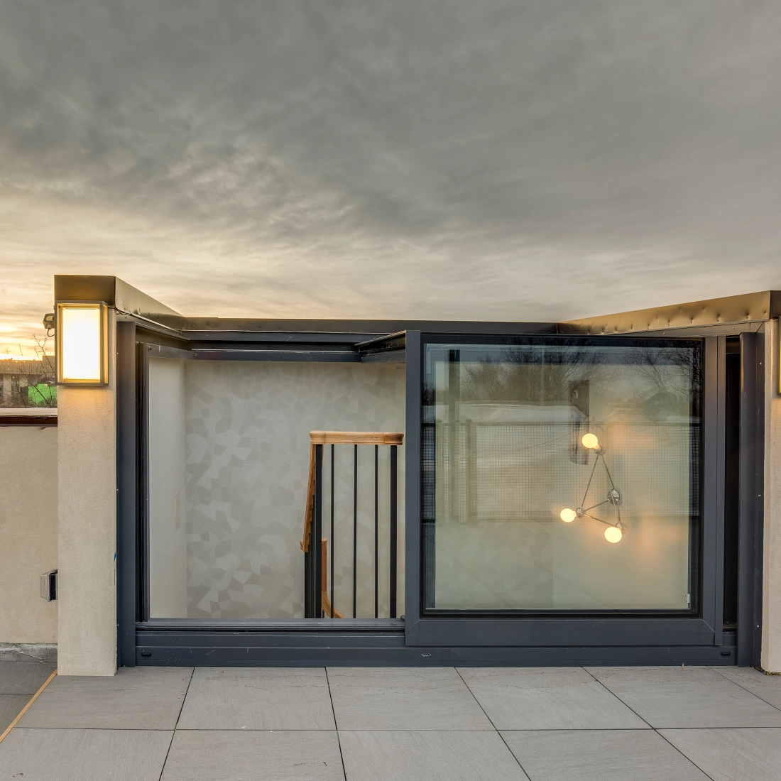 A modern rooftop terrace with large glass doors leading to an indoor space. Warm lighting fixtures flank the entrance, and a staircase is visible inside. The sky is cloudy with a hint of sunset in the background.