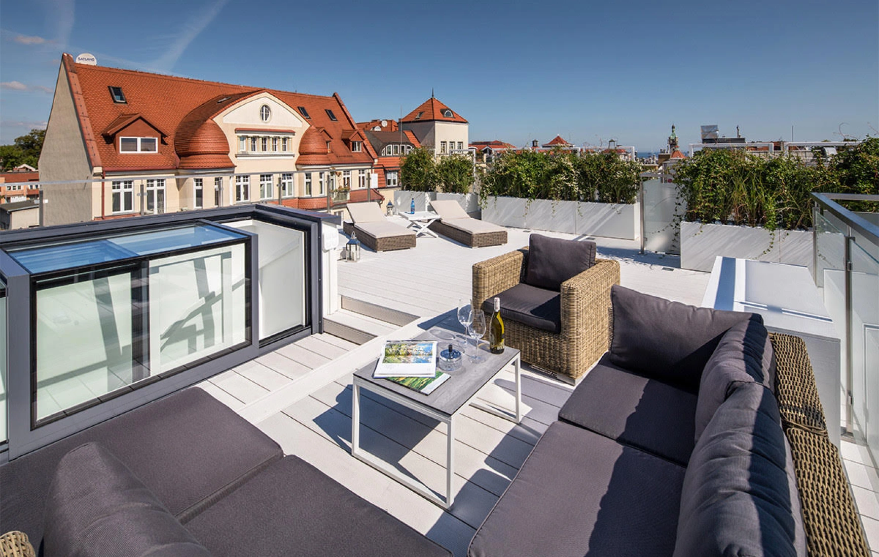 A stylish rooftop terrace features modern outdoor furniture, including a cozy sectional sofa and a small table. The space is surrounded by greenery, with a city skyline and a clear blue sky in the background.