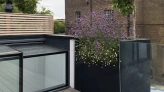 A rooftop terrace features a wooden deck and modern black and clear glass planters filled with purple flowers. In the background, a brick building and a partly cloudy sky are visible.