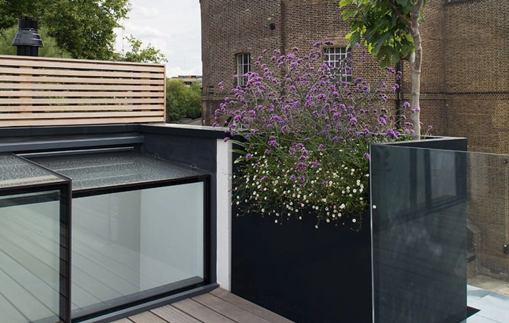 A rooftop terrace features a wooden deck and modern black and clear glass planters filled with purple flowers. In the background, a brick building and a partly cloudy sky are visible.