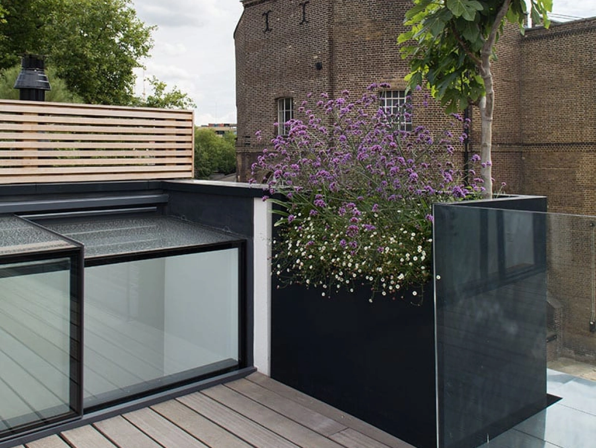 A modern rooftop terrace featuring wooden flooring and a large planter with purple flowers and greenery. In the background, a brick building with windows and a cloudy sky can be seen.