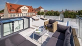 A stylish rooftop terrace features modern outdoor furniture, including a cozy sectional sofa and a small table. The space is surrounded by greenery, with a city skyline and a clear blue sky in the background.