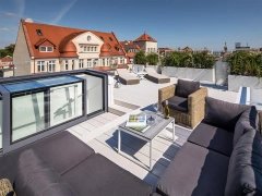 Terraza moderna con muebles de exterior cómodos, incluyendo un sofá y una mesa, con una vista clara del cielo y edificios cercanos. Se aprecian áreas ajardinadas y una piscina al fondo.