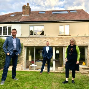 Three people stand in front of a stylish modern home with large windows. The house is two stories high and features a stone facade, surrounded by a green lawn.