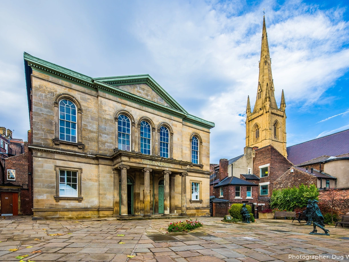 Historic stone building with columns stands in the foreground, surrounded by a paved area and flower beds. In the background, a tall church spire rises against a blue sky with clouds.