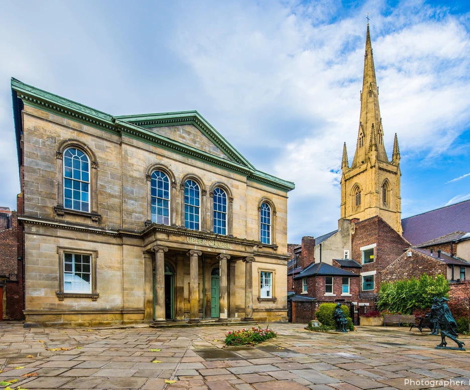 Historic stone building with columns stands in the foreground, surrounded by a paved area and flower beds. In the background, a tall church spire rises against a blue sky with clouds.